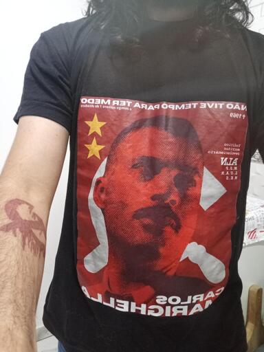 A imagem é uma "selfie" de espelho (o que faz com que os textos apareçam invertidos horizontalmente), mostrando o tronco e o braço esquerdo de uma pessoa de pele clara.
A Camiseta:
A pessoa veste uma camiseta preta com uma grande estampa retangular centralizada, em tons de vermelho e branco, com forte teor político e histórico:
Imagem Central: Um retrato estilizado em vermelho e preto de Carlos Marighella (escritor e ativista político brasileiro).
Símbolos: Atrás da cabeça dele, há uma grande foice e martelo brancas (símbolo comunista) e, no canto superior esquerdo, duas estrelas amarelas.
Texto no Topo: A frase célebre: "NÃO TIVE TEMPO PARA TER MEDO". Logo abaixo, em letras menores: "o inimigo número 1 da ditadura".
Texto na Lateral Direita: Uma lista com as palavras "político", "escritor", "revolucionário", a sigla "ALN" e o ano "1969" acompanhado de uma cruz.
Texto na Base: O nome "CARLOS MARIGHELLA".
Detalhes Físicos:
Cabelo: A pessoa tem cabelos longos, escuros e ondulados que caem sobre os ombros.
Tatuagem: No antebraço esquerdo exposto, há uma tatuagem em tinta vermelha (ou cicatrizada/escarificada)