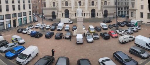 Foto panoramica di piazza Affari a Milano. Tutta la piazza è occupata da auto parcheggiate in maniera illegale. In mezzo  alla piazza, circondato da auto, c'è il famoso "dito" di Cattelan. Alcuni turisti si stanno facendo una foto con il monumento in mezzo alle auto parcheggiate.